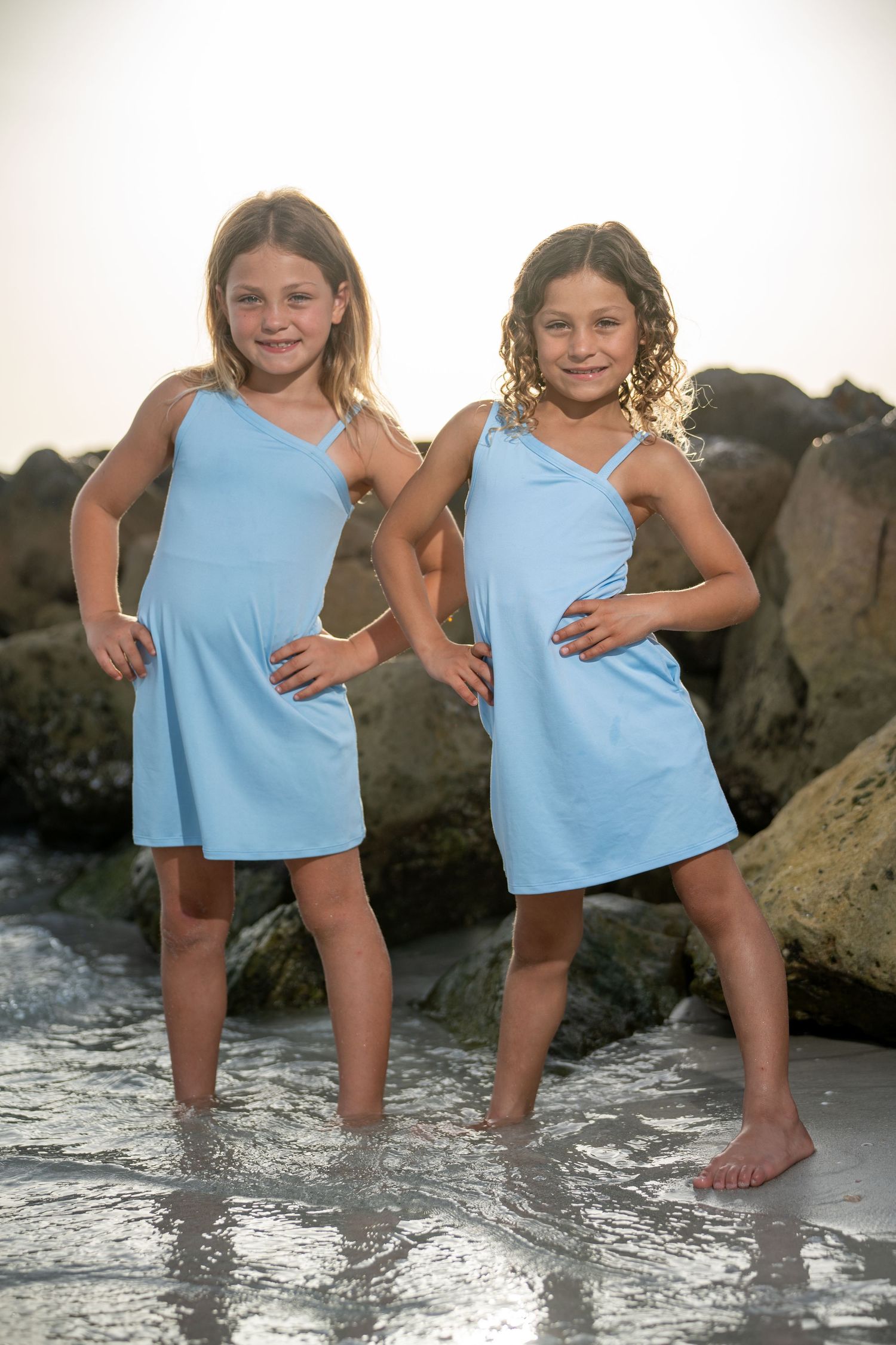 Two children in matching light blue dresses stand playfully in shallow water near rocky beach at sunset.