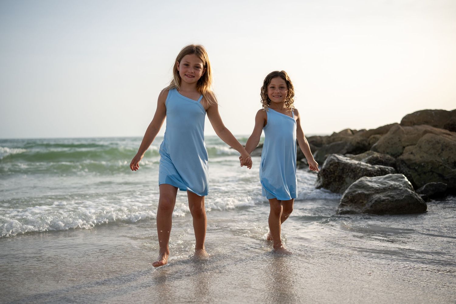 Two people in matching blue dresses walk hand in hand along a beach at sunset near rocky outcrops and crashing waves.