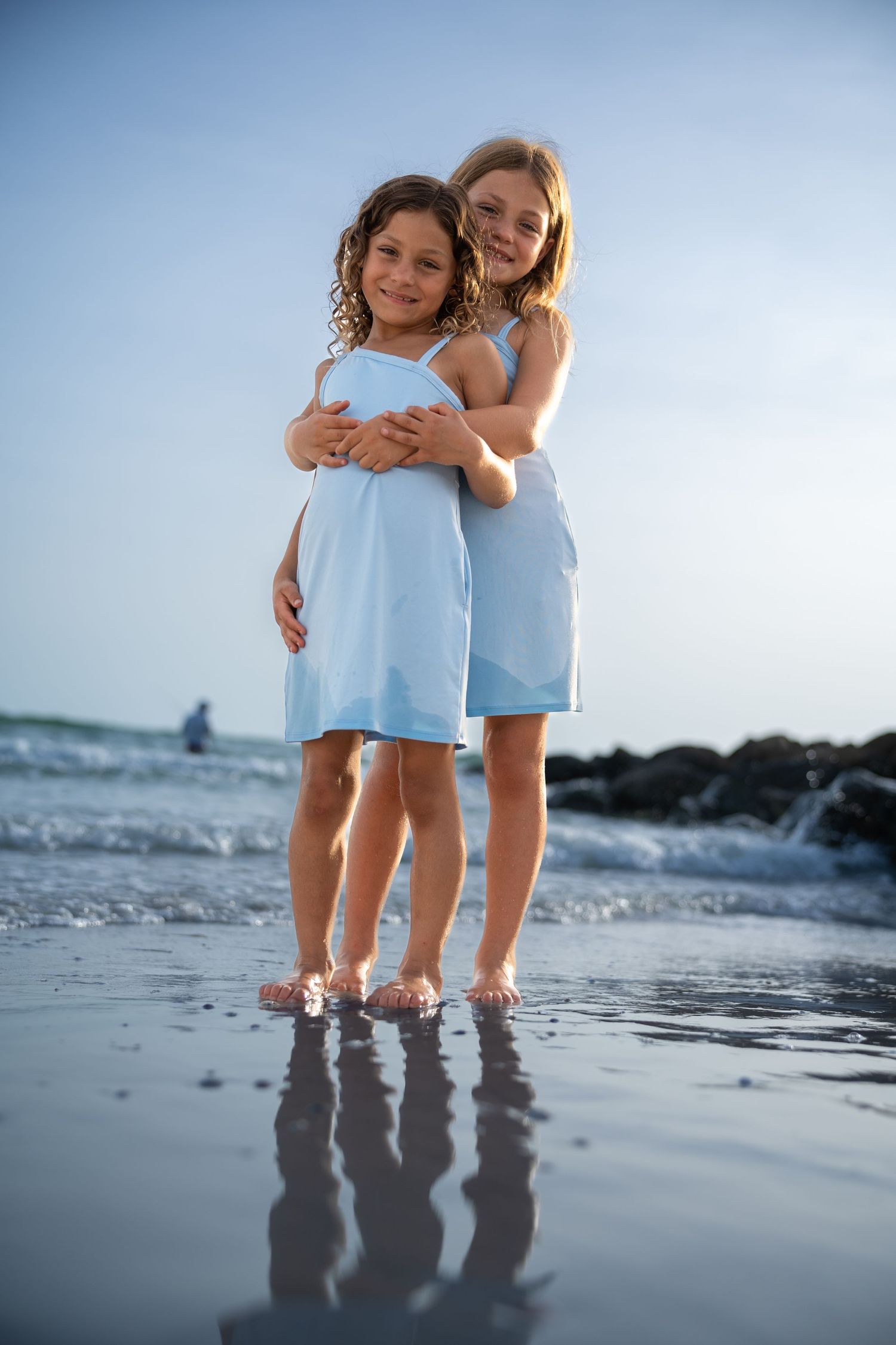 Two children in light blue dresses hug while standing at the edge of the ocean waves on a sandy beach at sunset.