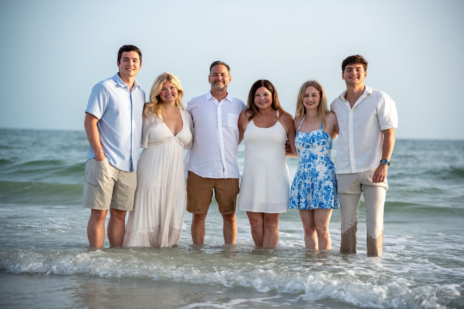 A group of people wearing white and blue outfits stand together at the ocean's edge during sunset.