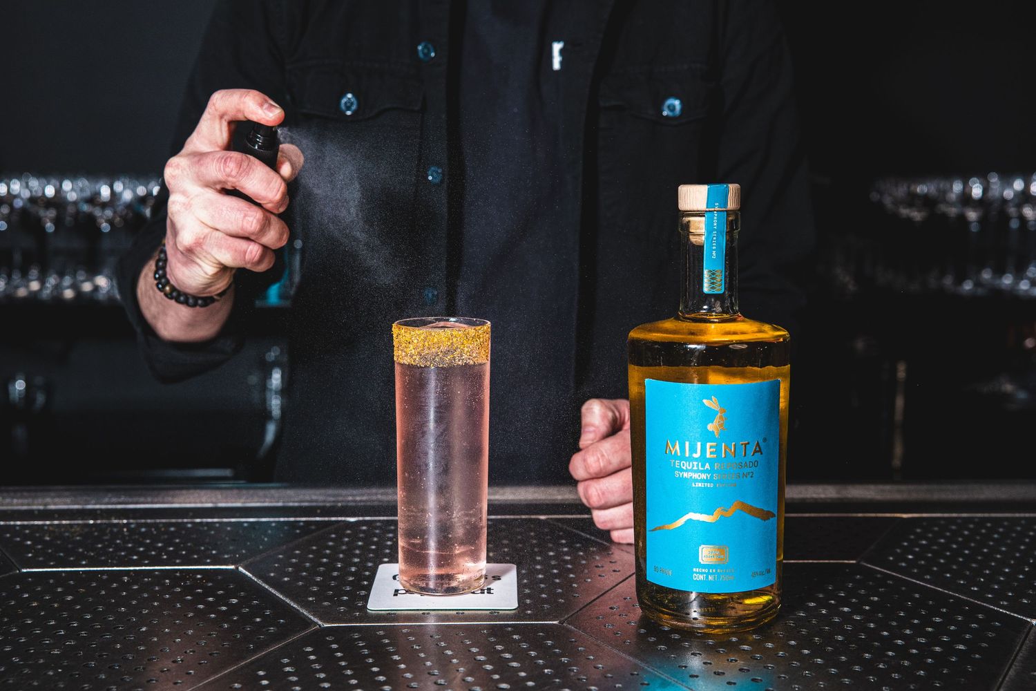 A bartender pours a crafted cocktail next to a blue-labeled bottle on a dark bar counter.