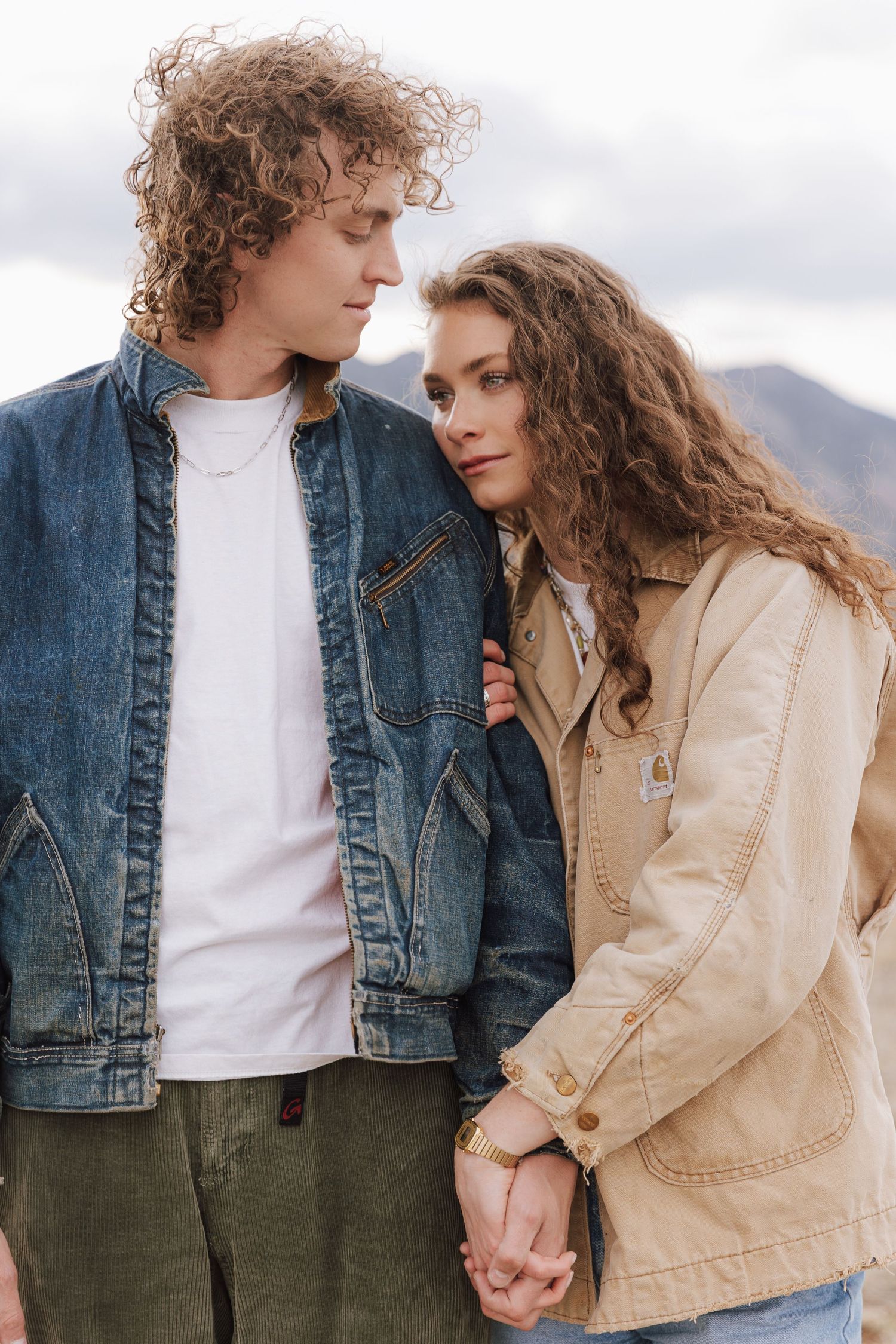 Two people in casual denim and beige jackets hold hands and share intimate moments outdoors against a cloudy backdrop.