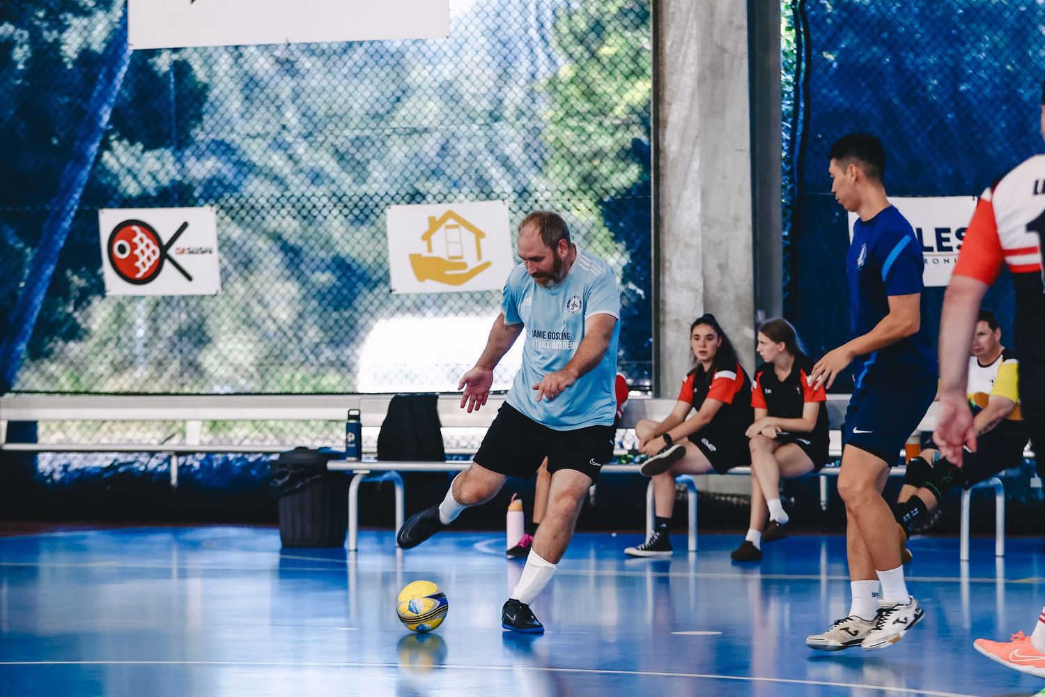 People play indoor soccer on a large white floor in a spacious indoor facility with natural lighting.