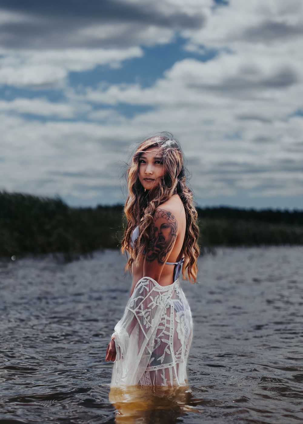 Woman in sheer white dress standing in lake water under cloudy sky.