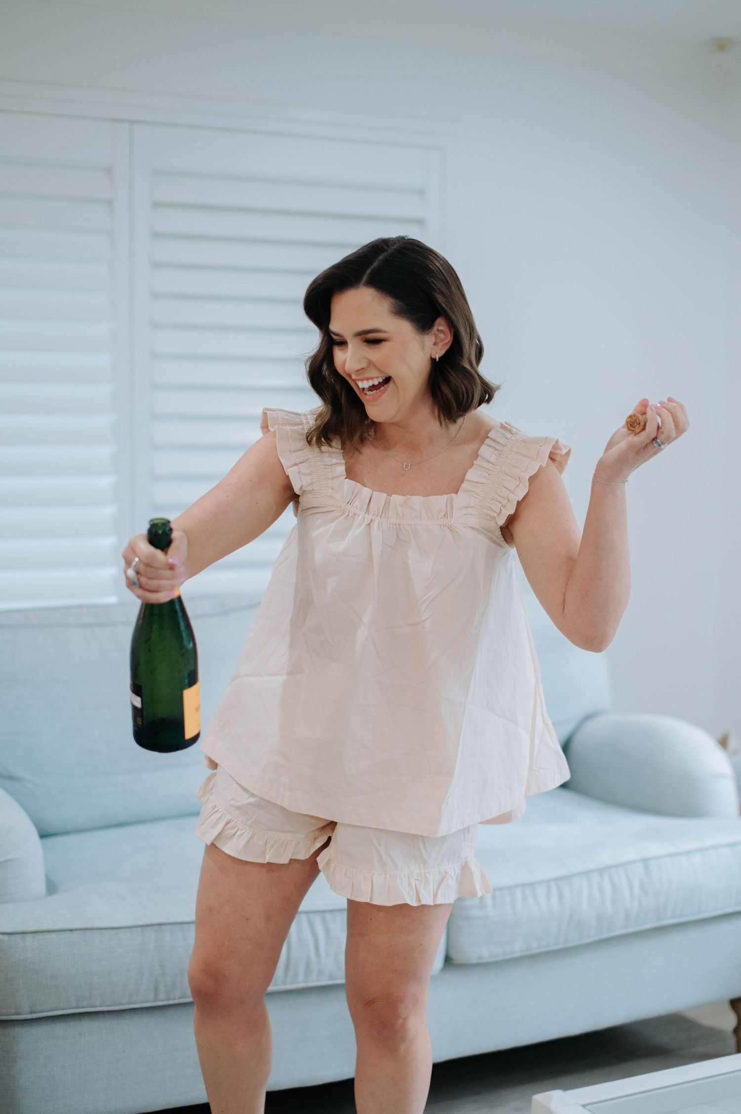 Woman in light pink ruffle pajama set dances joyfully while holding champagne bottle in bright modern room.