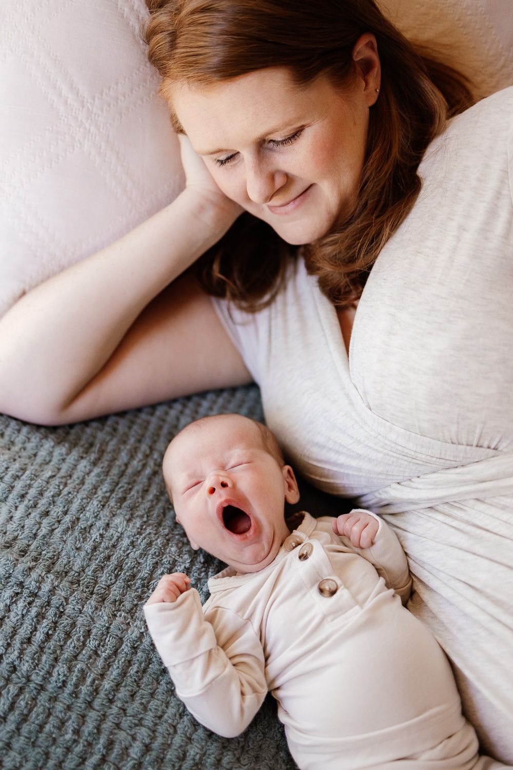 Mom lays next to her yawning sleepy baby on their bed during their in-home newborn photos