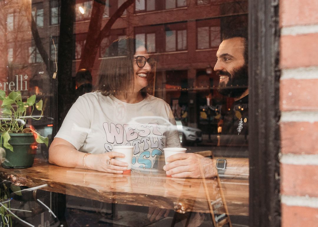 Through a window reflection, two friends share drinks at a cozy brick-walled cafe.