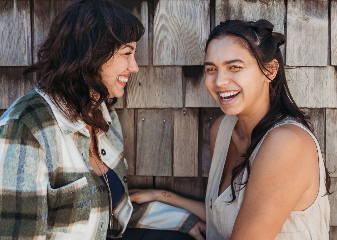Two friends sharing a joyful moment of laughter while sitting together against a wooden wall.