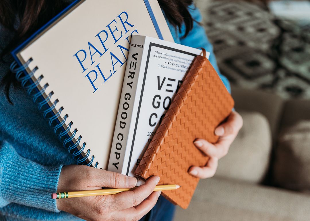 Person in blue sweater holding multiple notebooks including Paper Planner and lined notebooks with orange cover.