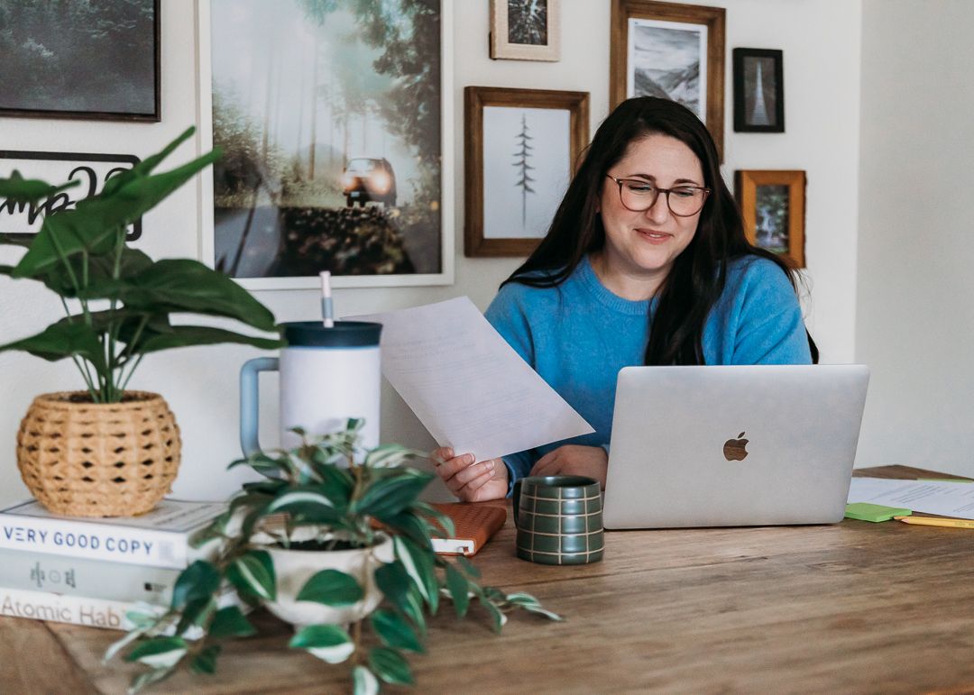 A person in a blue shirt sits at a wooden desk working on a MacBook laptop surrounded by indoor plants and framed artwork.