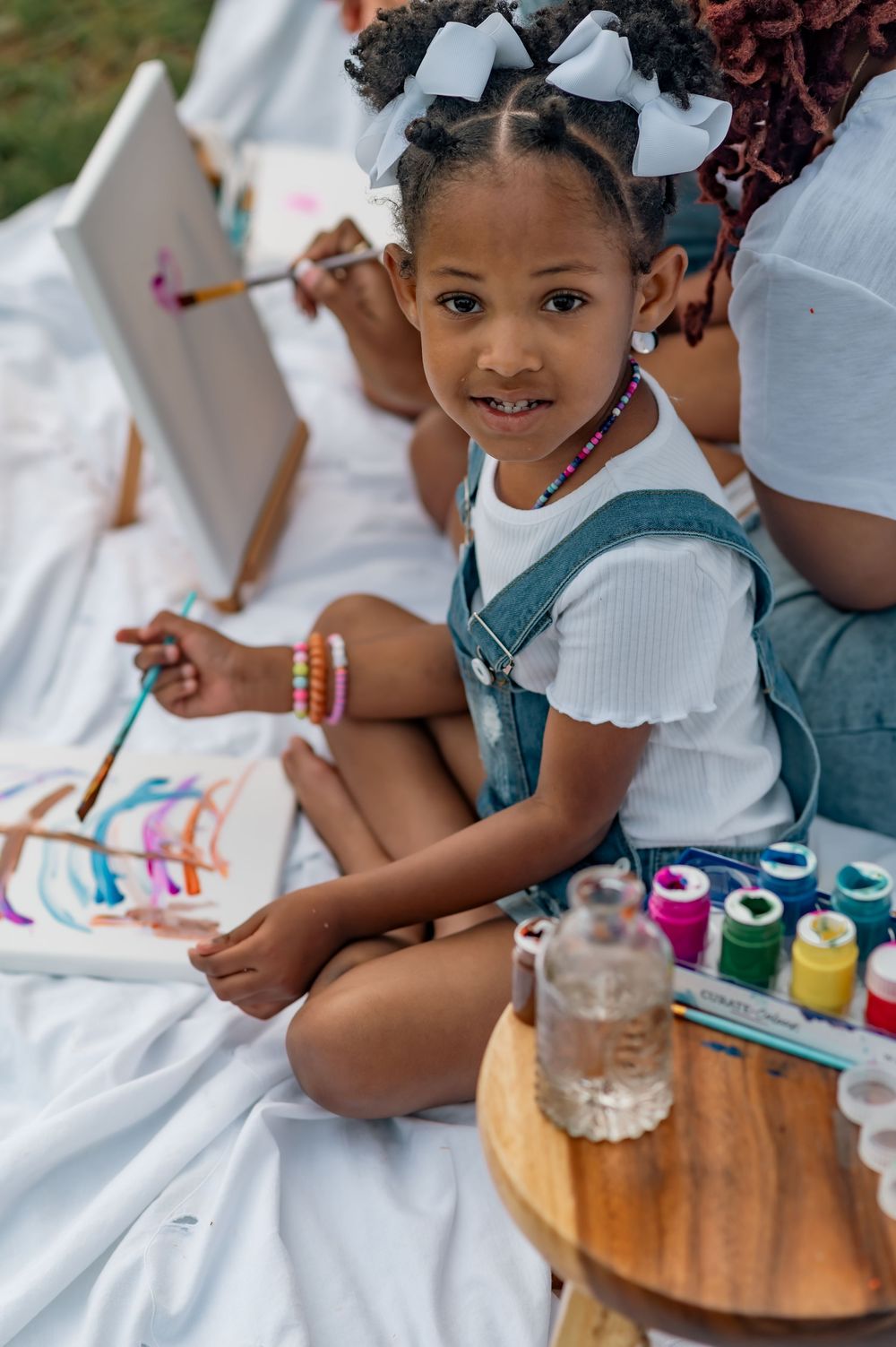 Young artist sits on white blanket enjoying outdoor painting session with colorful paints and art supplies.