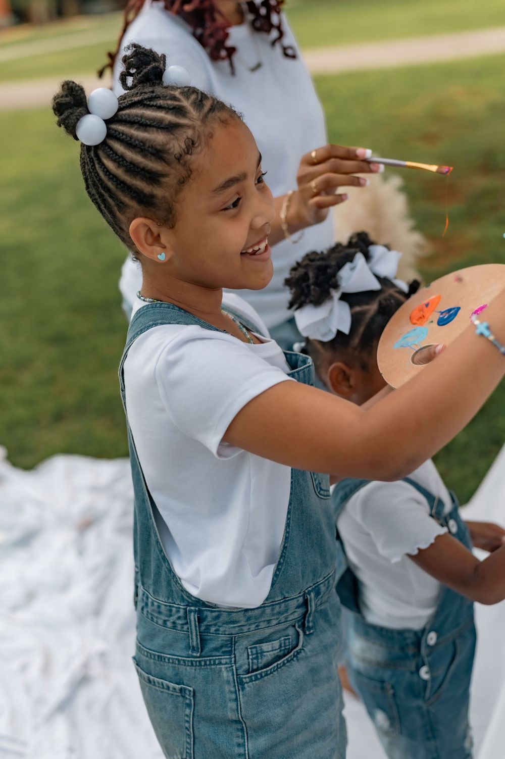 Child in denim overalls and white shirt smiling while holding a painting brush outdoors in a park setting.