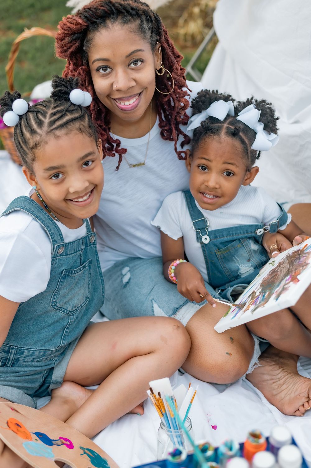Family enjoys creative art time together on white blanket with painting supplies and denim overalls.