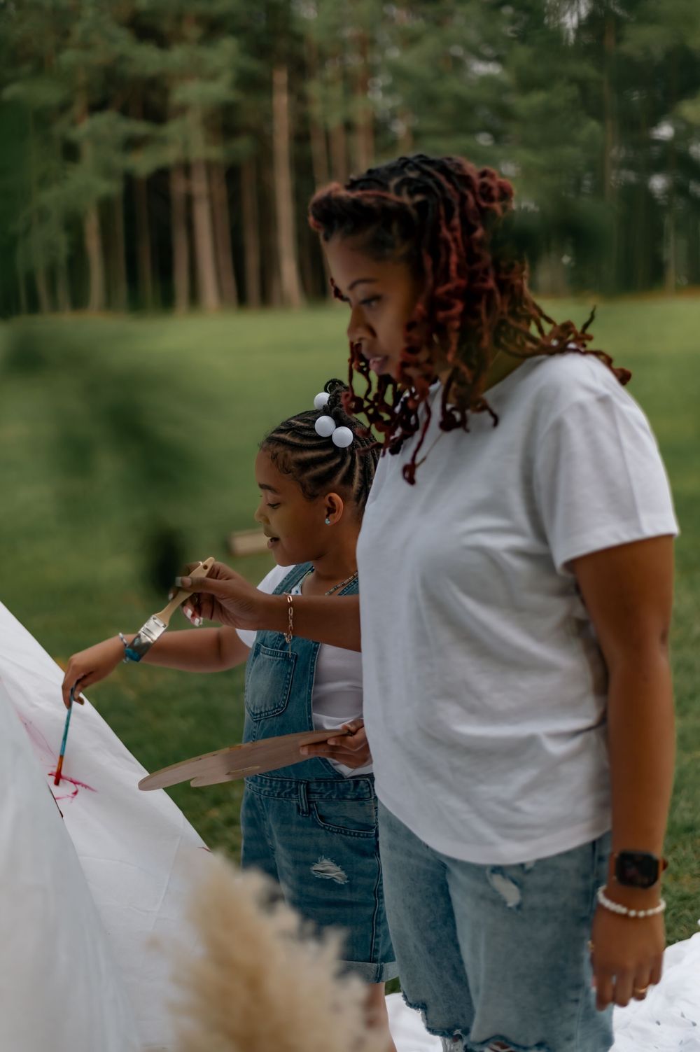 Two people in casual wear painting together on a canvas outdoors in a pine forest setting.