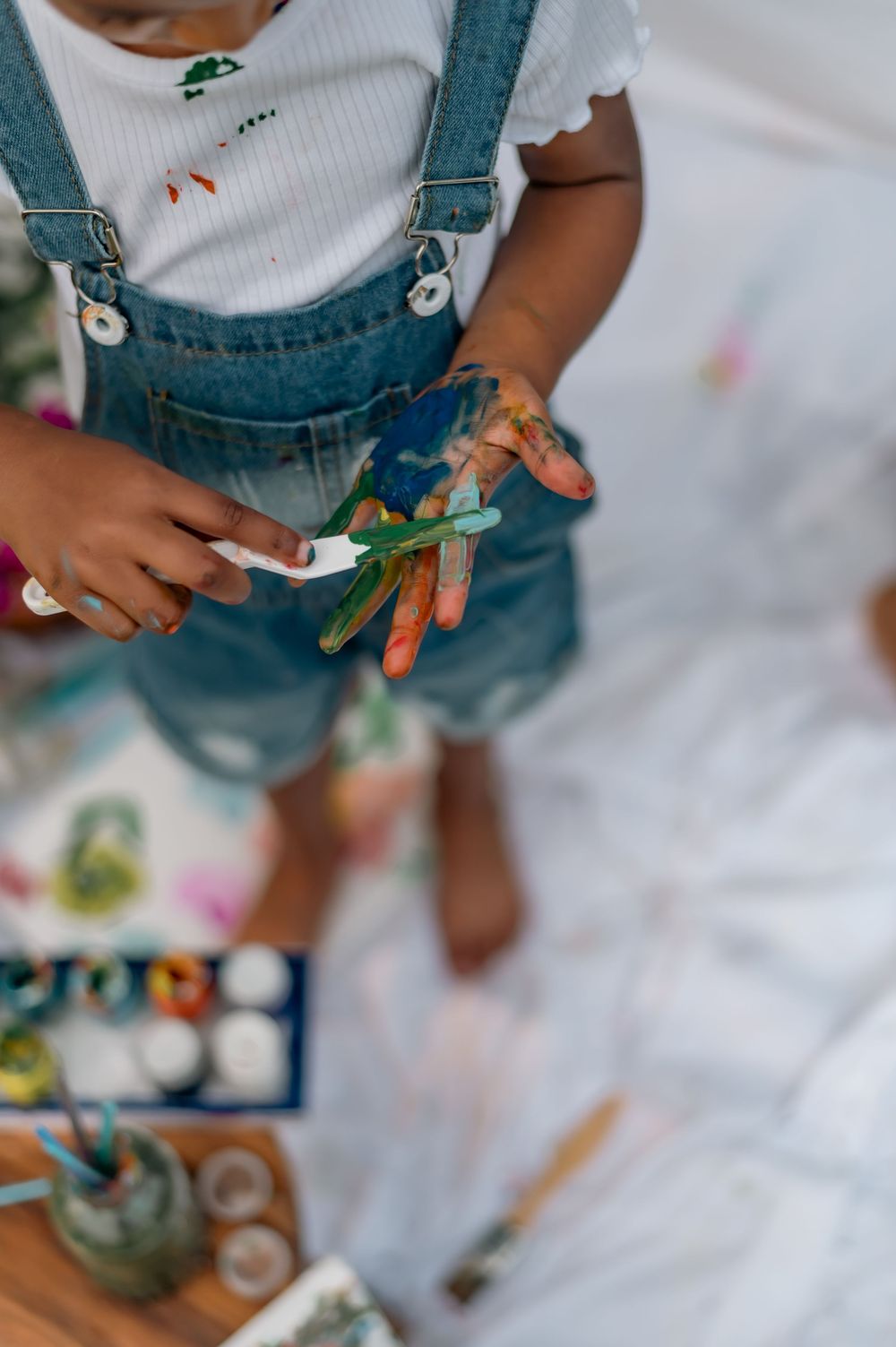 Child in denim overalls holds colorful art supplies while creating artwork on a white surface during playtime.