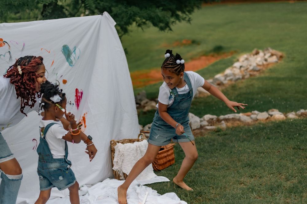 Children playing and running past a white sheet backdrop in an outdoor garden setting with colorful paint splatters.