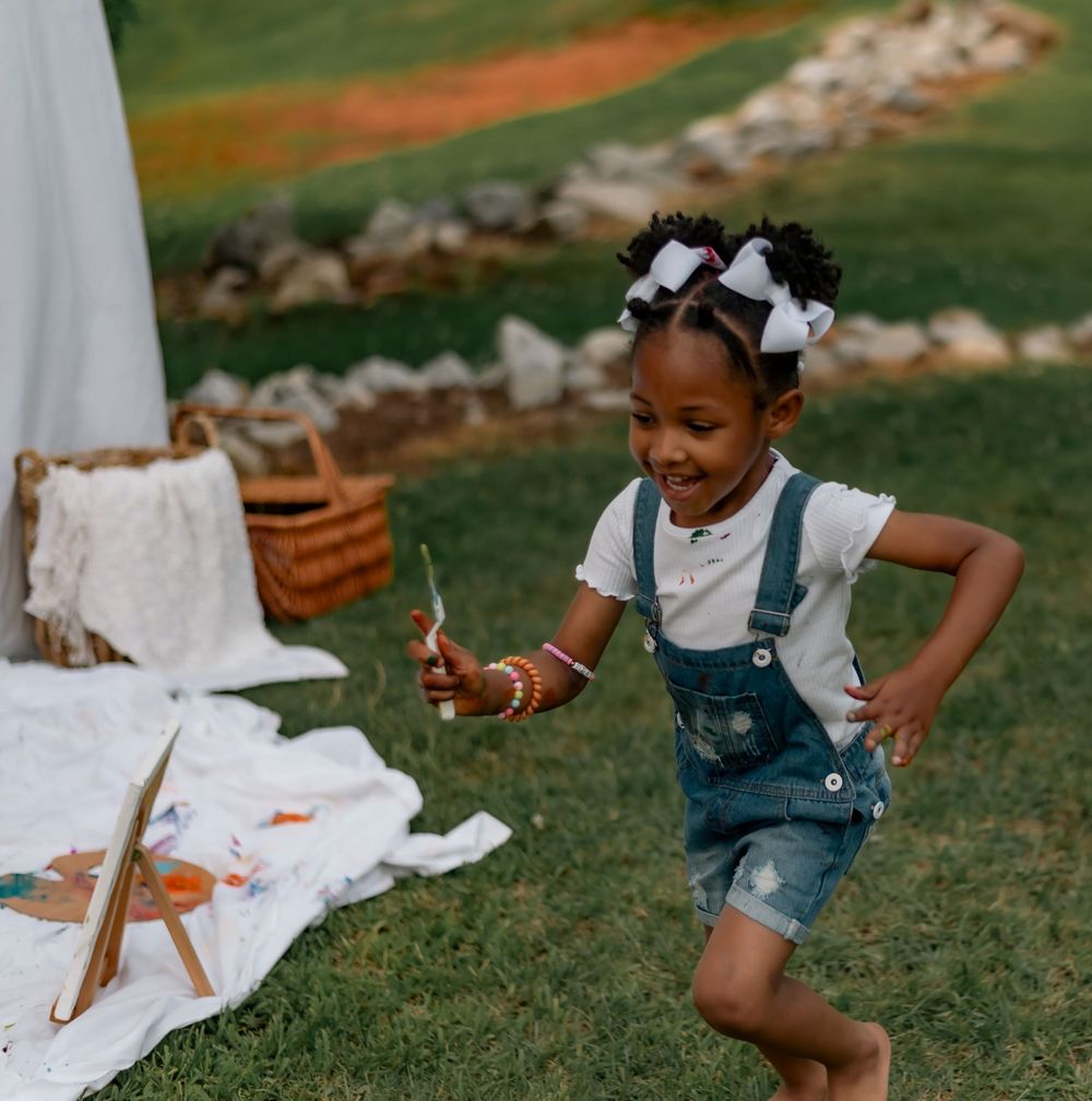 Child in denim overalls plays outdoors on grass, reaching down toward something on a white blanket or sheet.