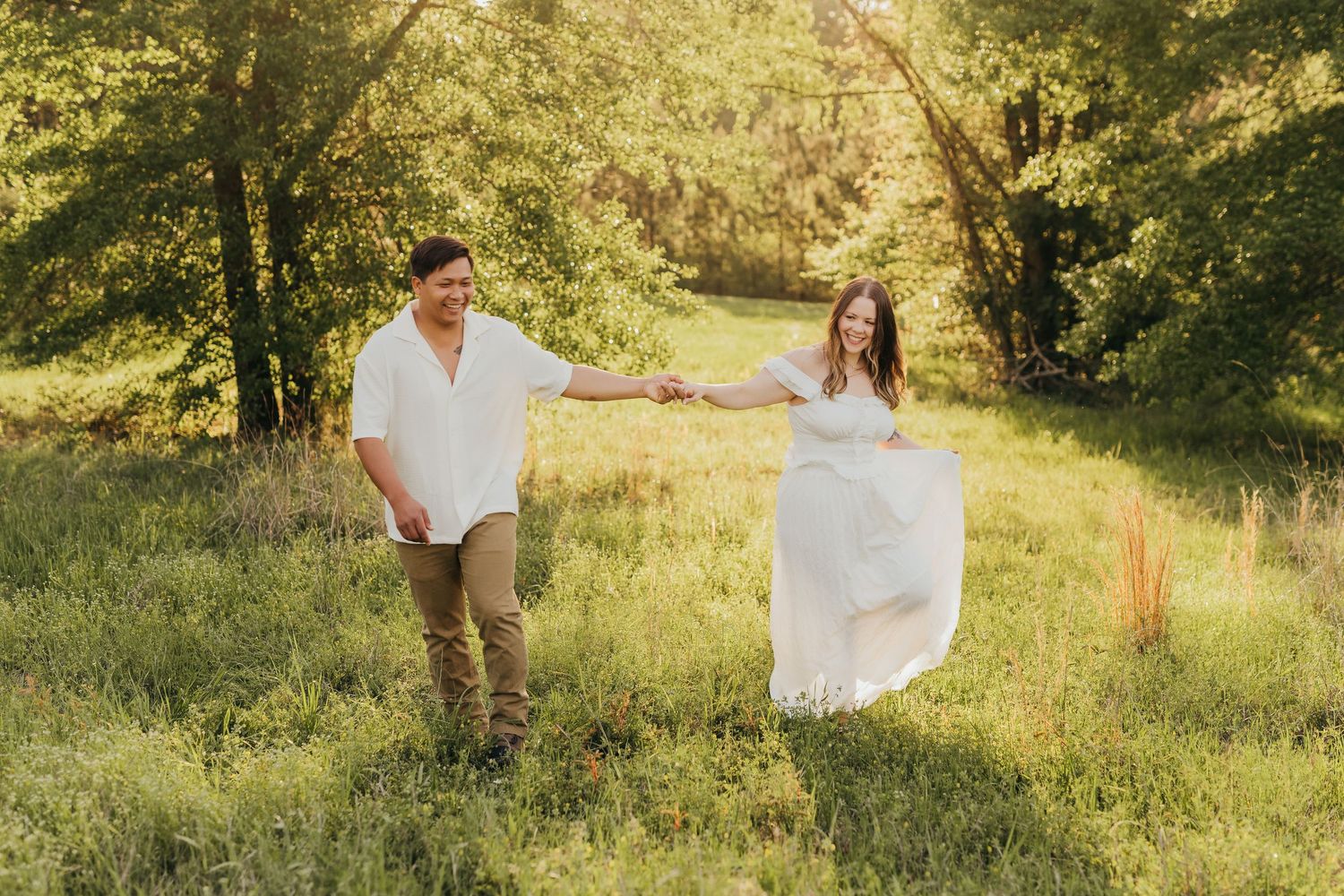 Two people stand in a sunlit pasture with green grass and trees during a romantic outdoor photo session in Tyler, Tx.