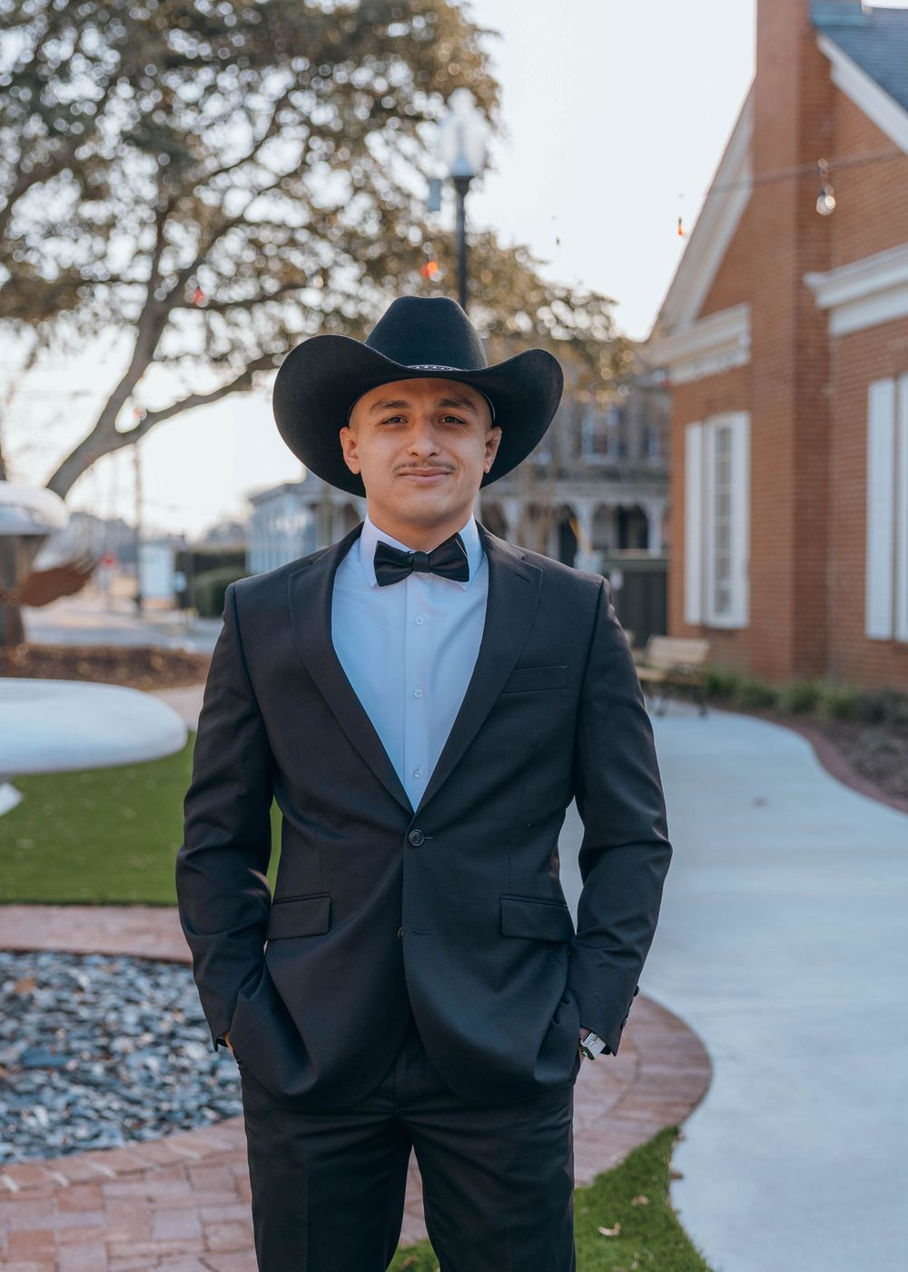 A person in a black suit and cowboy hat stands in front of a brick building on a sunny day. Groom. North Carolina.