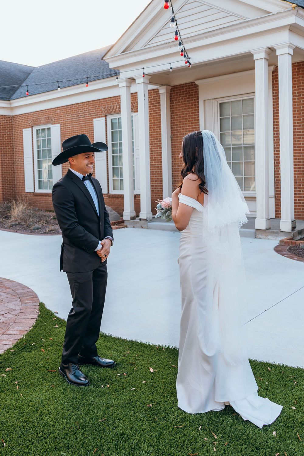 A couple in wedding attire playfully poses with a flowing veil outside a brick building. First look. Wedding day. North Carolina.