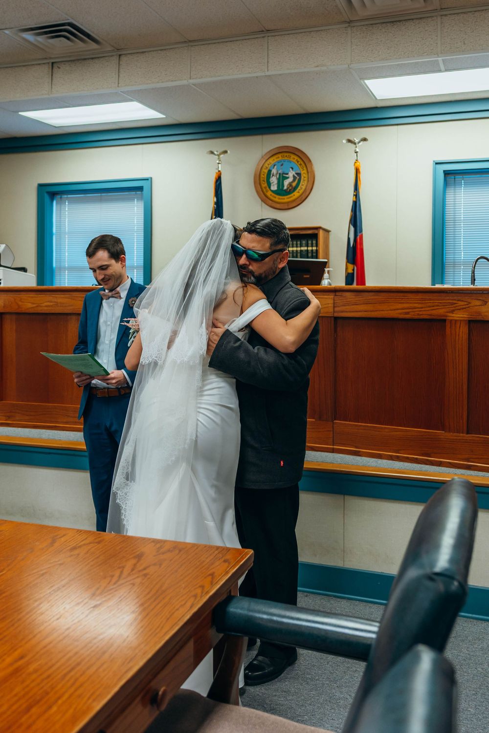 A wedding ceremony moment in a courthouse with turquoise trim and wooden furnishings. Father daughter hug.
