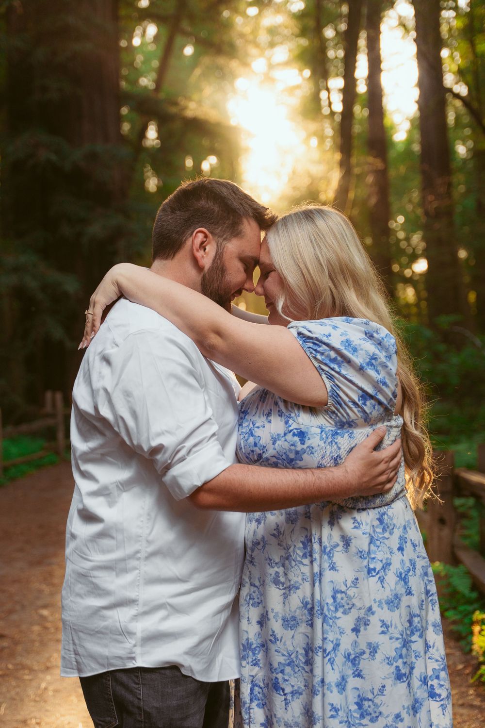 Romantic couple embraces on forest path with sunlight streaming through trees during golden hour.