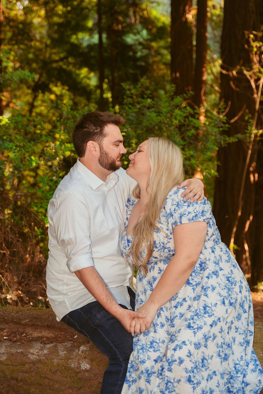 A couple in coordinating white and blue outfits share an intimate moment in a sunlit forest setting.
