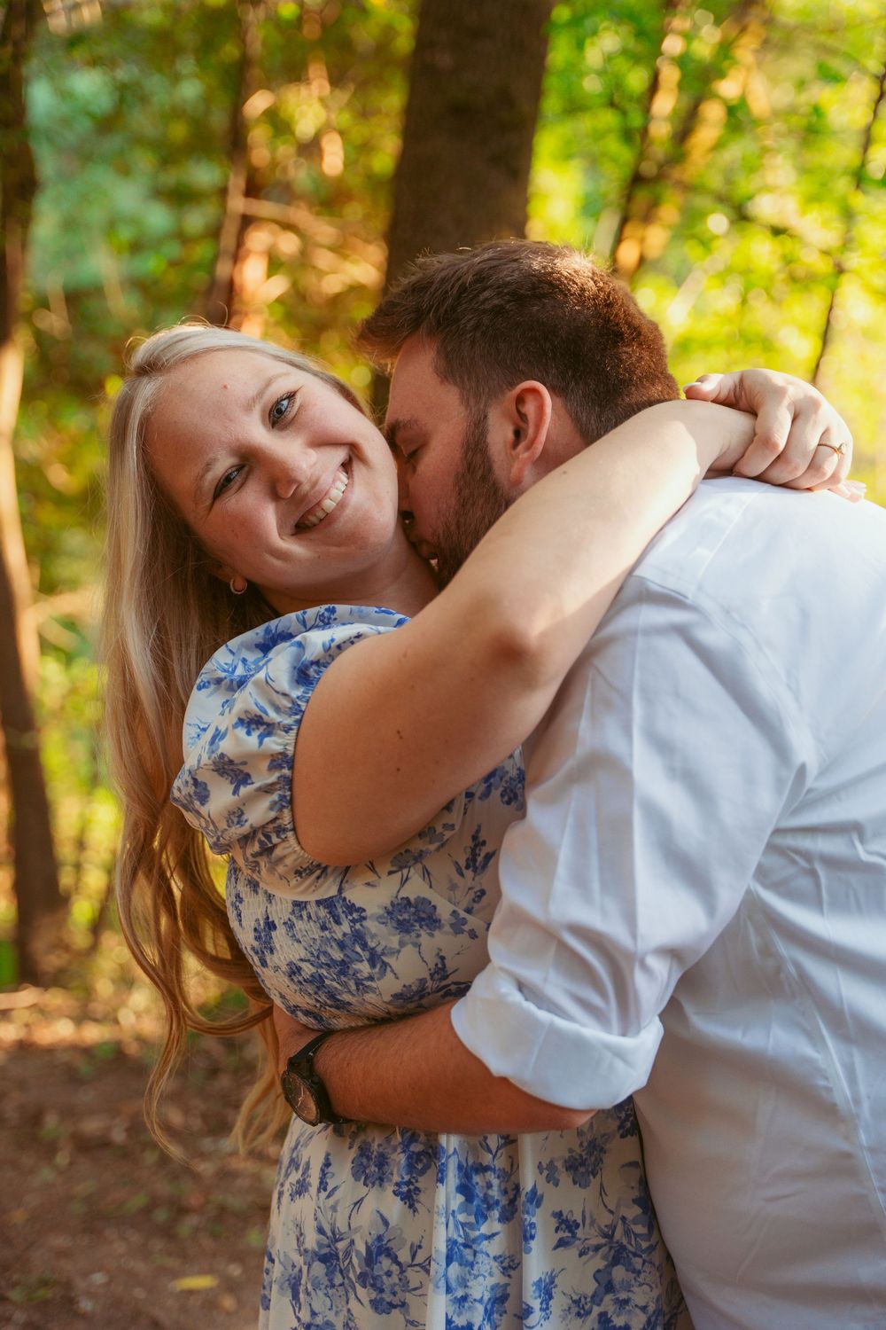 Couple embraces in romantic moment among trees with sunlit forest backdrop during outdoor photoshoot.