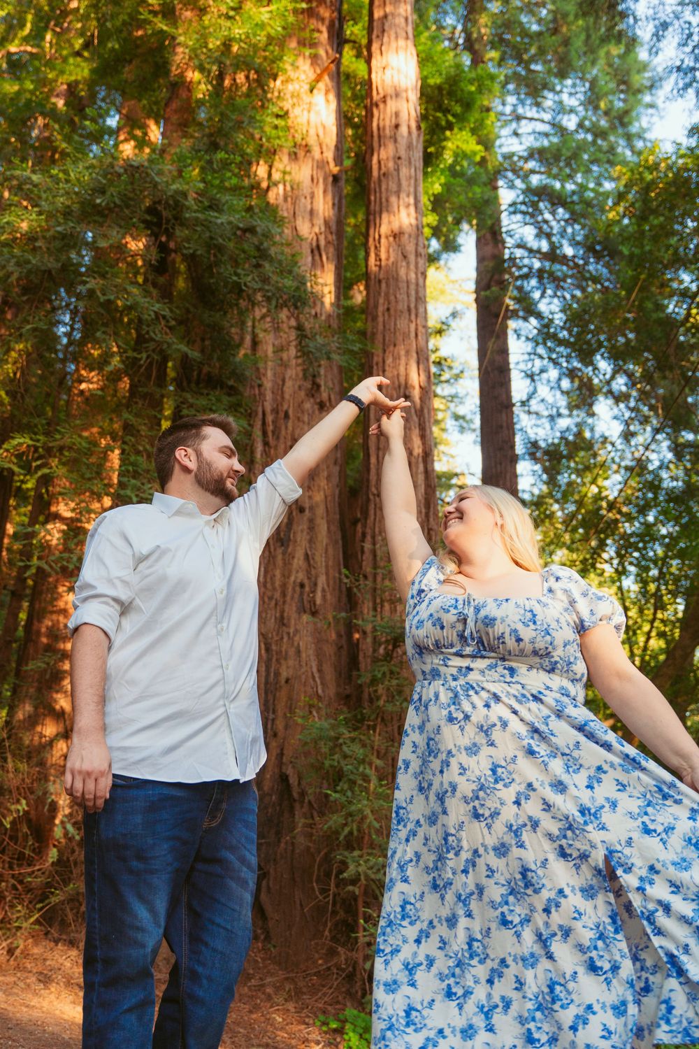 A couple dancing together on a forest trail surrounded by towering redwood trees during golden hour.