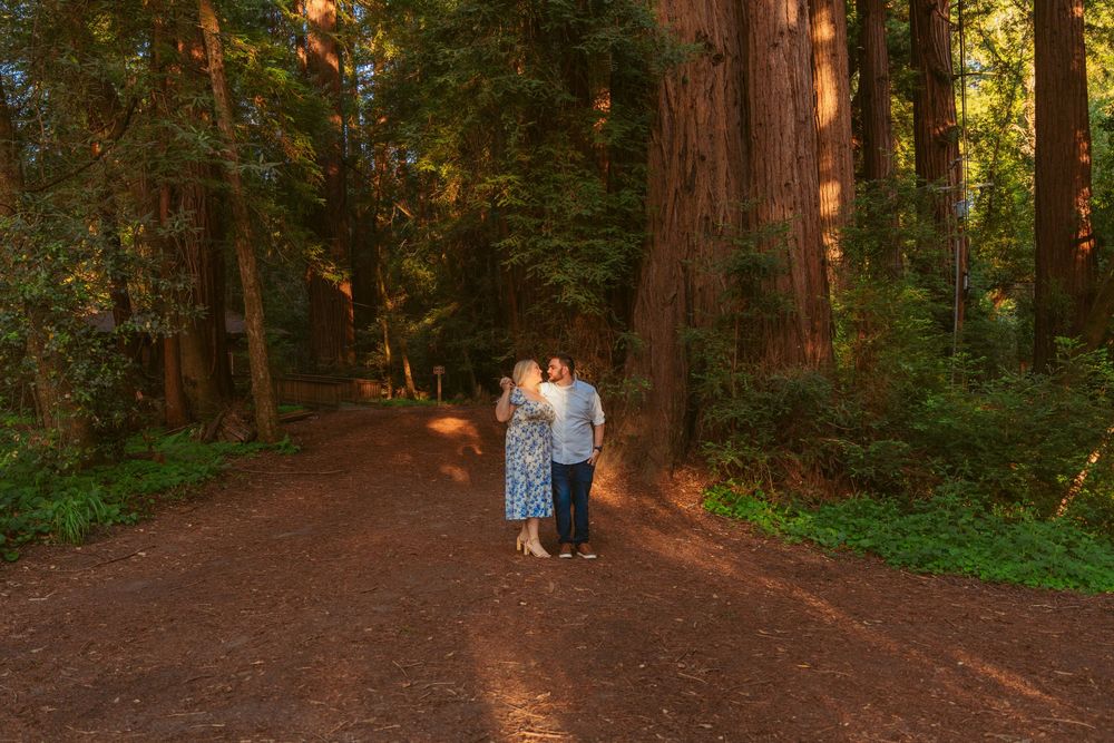 Two people share an intimate moment while walking along a sunlit forest trail lined with towering redwood trees at sunset.