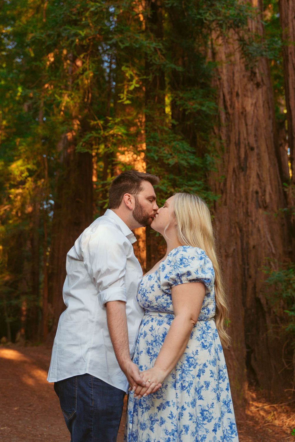 Romantic couple in white and blue floral dress standing together in a sunlit redwood forest.