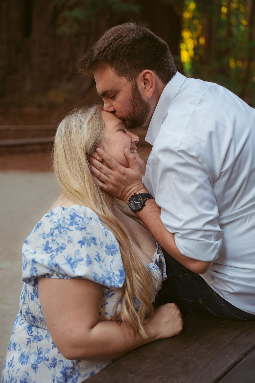 A tender romantic moment shared between a couple at a rustic wooden picnic table in a forest setting.