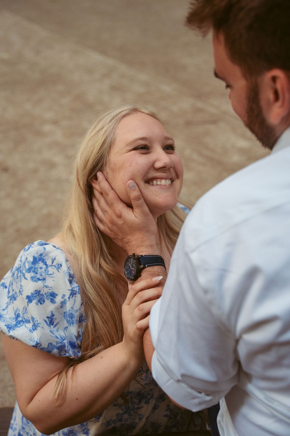 Happy couple sharing a tender moment as they embrace outdoors, with her in a blue floral dress and him in white shirt.