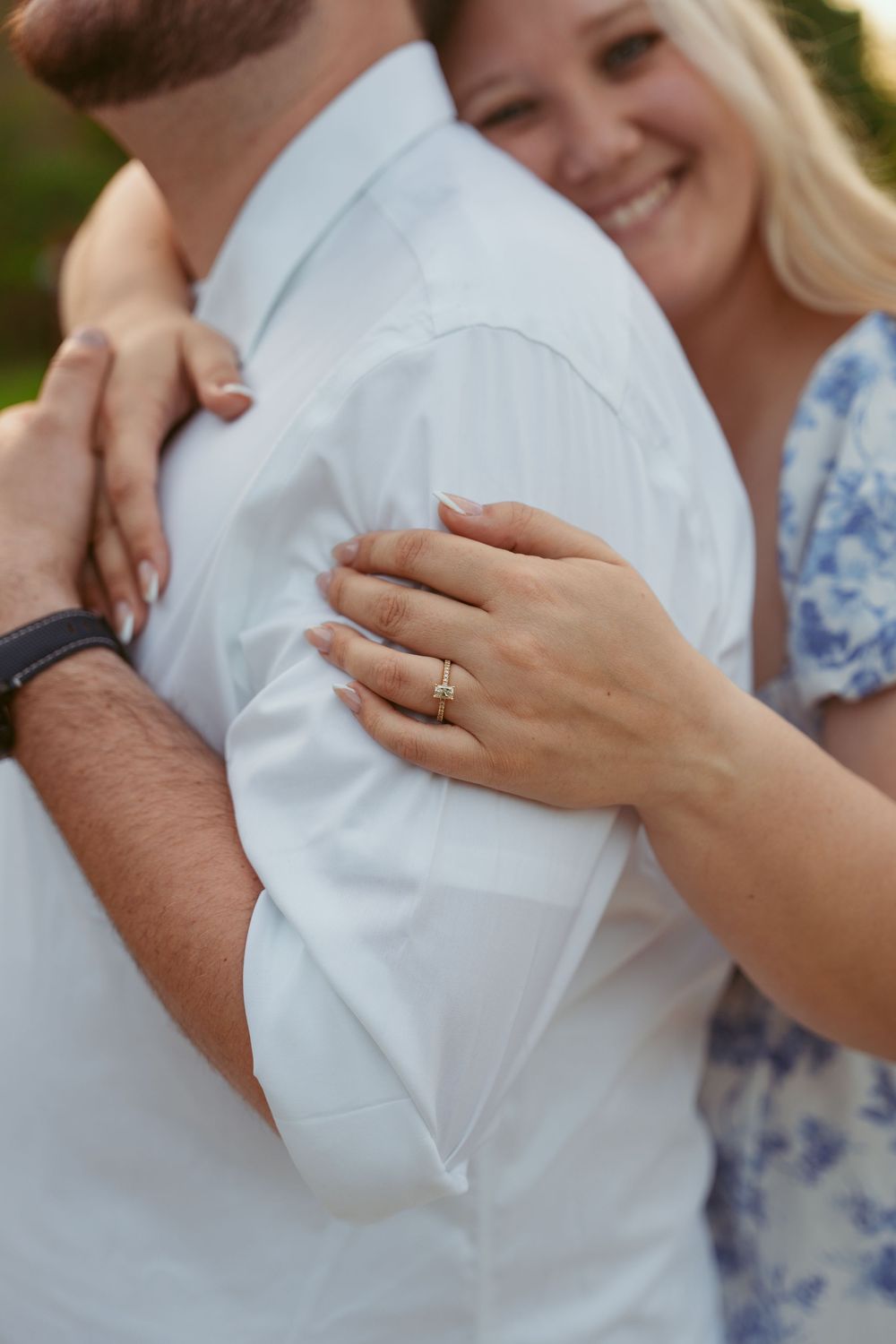 Close-up engagement photo showing hands embracing with diamond ring visible on blue and white floral dress.