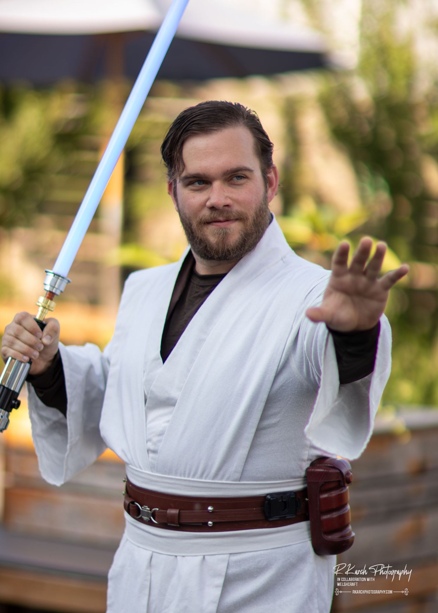 Person in white Jedi robes poses with a blue lightsaber while cosplaying as a Star Wars character in an outdoor setting.
