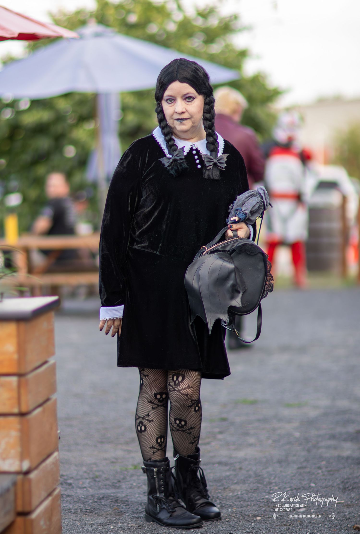 Person dressed as Wednesday Addams in a black velvet dress with braids and patterned tights poses outdoors.
