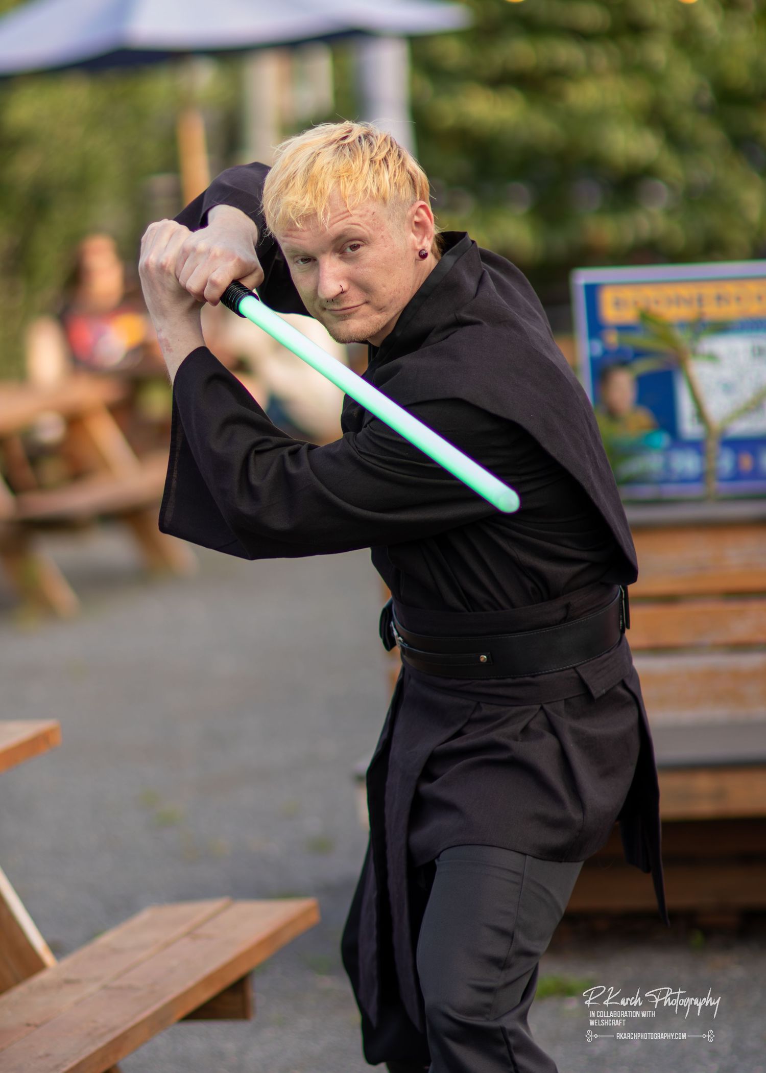 A figure in a black hooded costume poses with a glowing turquoise lightsaber in an outdoor setting.