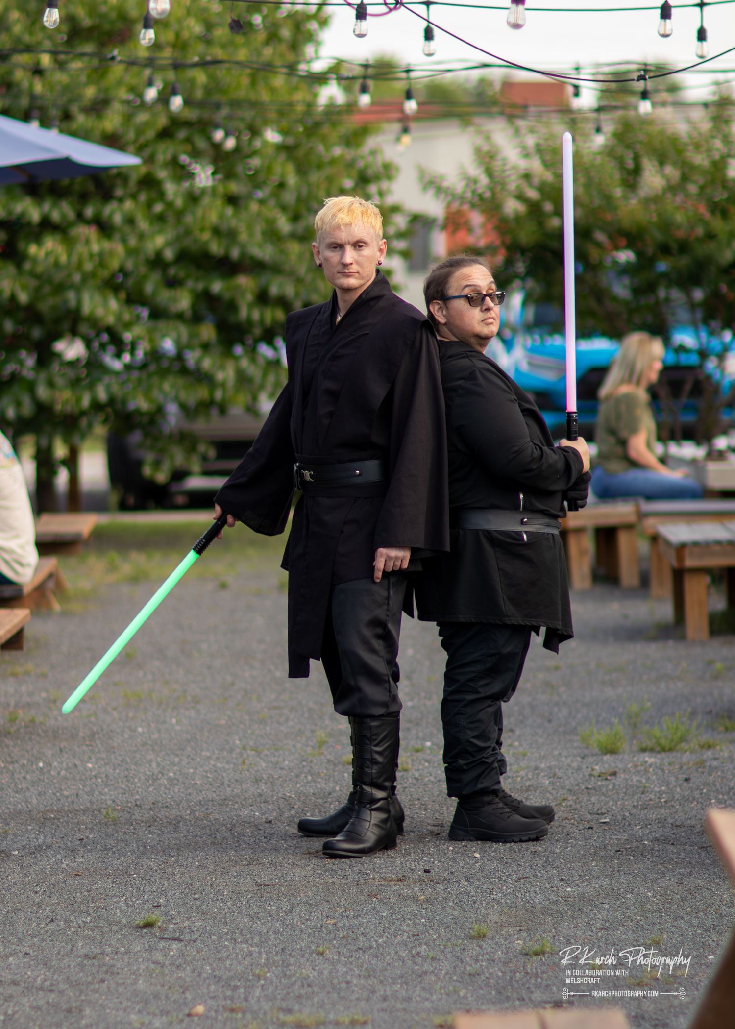 Two cosplayers in black outfits pose back-to-back at an outdoor event, one holding a green lightsaber prop.