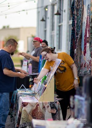 Vendor displays items at outdoor market booth while customer browses merchandise.