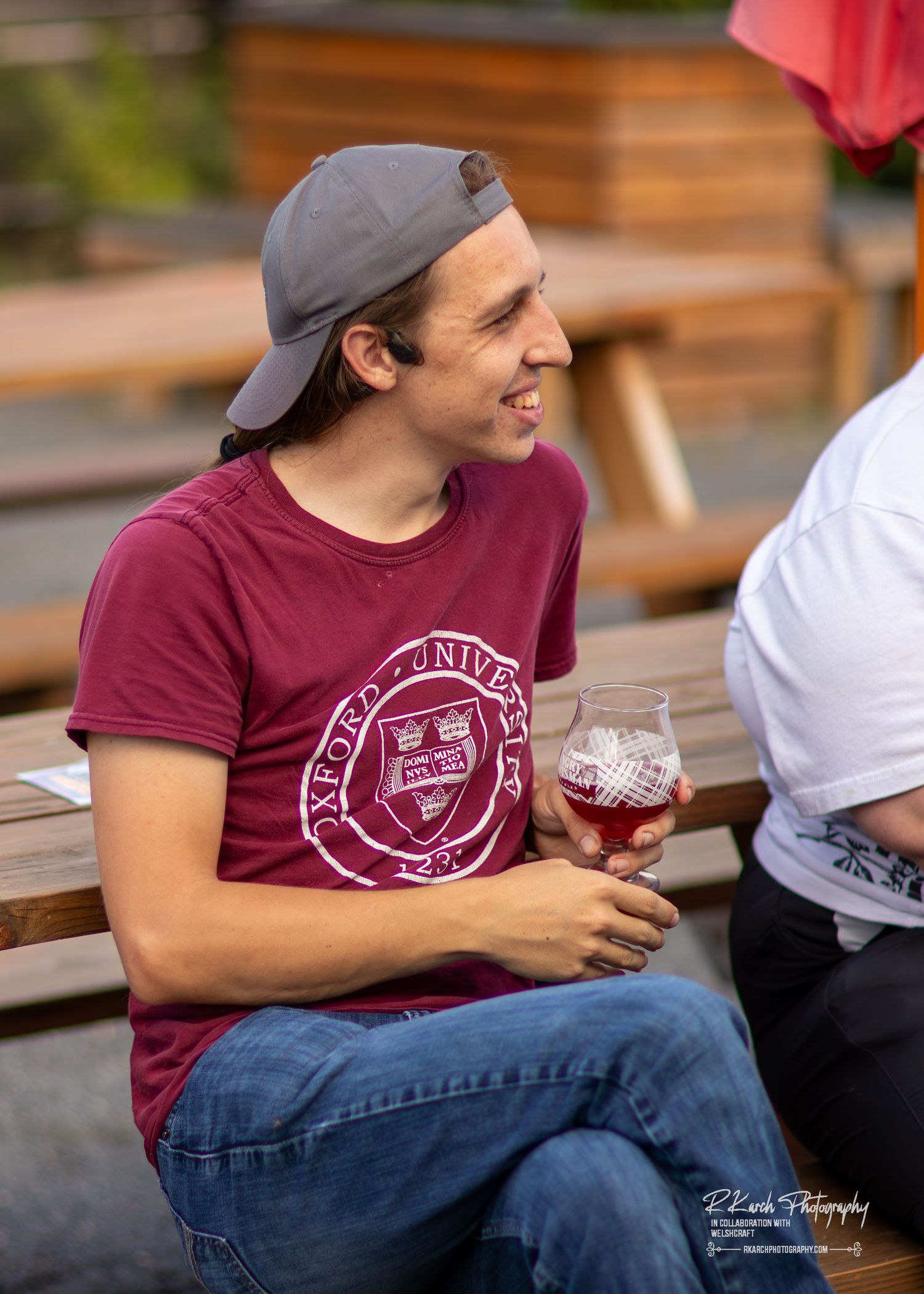 Person wearing college logo t-shirt and baseball cap smiling while holding drink at outdoor gathering.