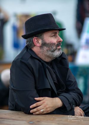 Person wearing a black fedora and dark jacket sitting at wooden table outdoors.