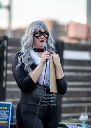 A cosplayer in a black corset and silver wig performs on stage with a microphone under string lights.