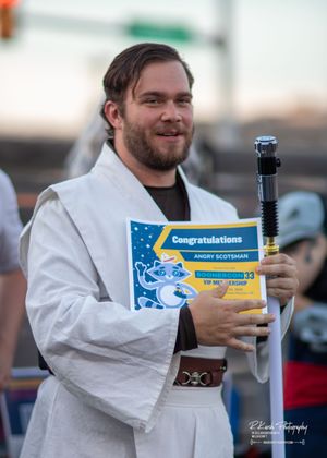 Person in white robe holds lightsaber prop and certificate at outdoor event.