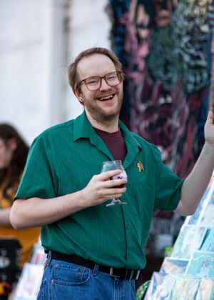 A person in a green shirt and glasses smiles while holding a drink at an outdoor social gathering.