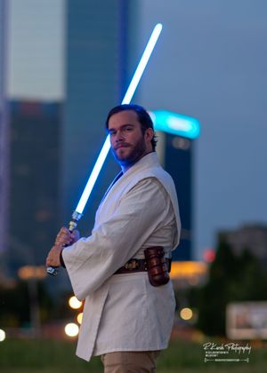 A person dressed in white Jedi robes poses with a glowing blue lightsaber against an evening cityscape.