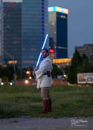 A lightsaber-wielding figure in Jedi-inspired costume stands in a grassy field at dusk with city buildings in the background.