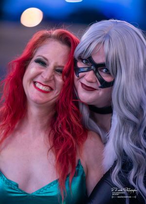 Two people with colorful dyed hair - red and silver - smile together wearing cosplay outfits against a blurred background.