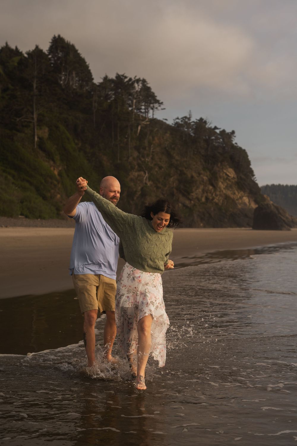 Two people dance together at the edge of the ocean with dramatic cliffs in the background at sunset.