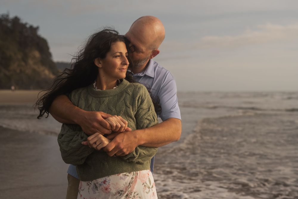 A couple embraces while standing at sunset on a peaceful beach with waves and cliffs in the background.