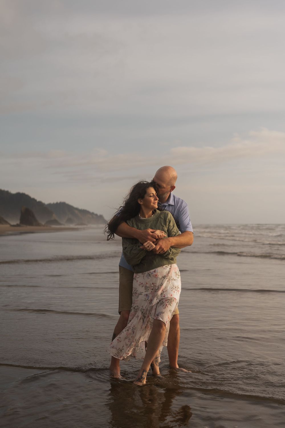 A romantic couple embraces while standing in the shallow ocean waves at sunset along a scenic coastline.